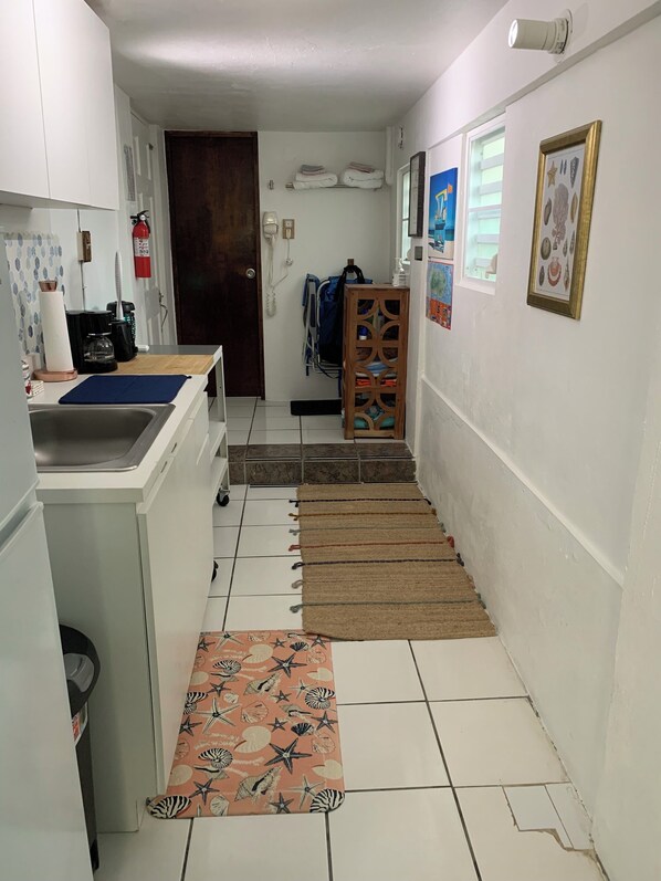 A narrow hallway with a kitchen area on the left, featuring a sink, cabinets, and a coffee machine. A wooden door and a fire extinguisher are visible on the wall. The hallway has a tiled floor with a rug leading to a staircase.