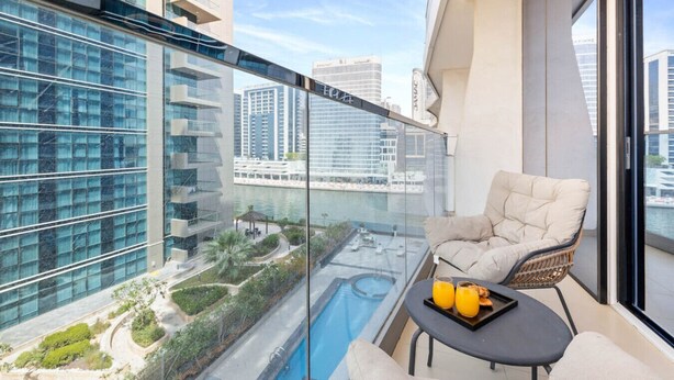 Uma varanda com guarda-corpo de vidro, uma cadeira estofada, uma pequena mesa com uma bandeja de laranjas e vista para uma piscina e edifícios ao redor.