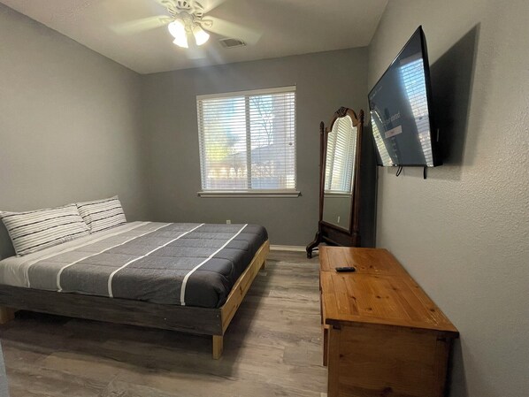 A bedroom with a bed, a wooden nightstand, a TV mounted on the wall, and a window with blinds.