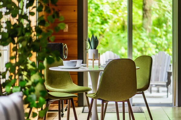 A dining area with a round table, four chairs, a potted plant, and a view of greenery outside. - The Copper Bird Cabin (Gatlinburg)