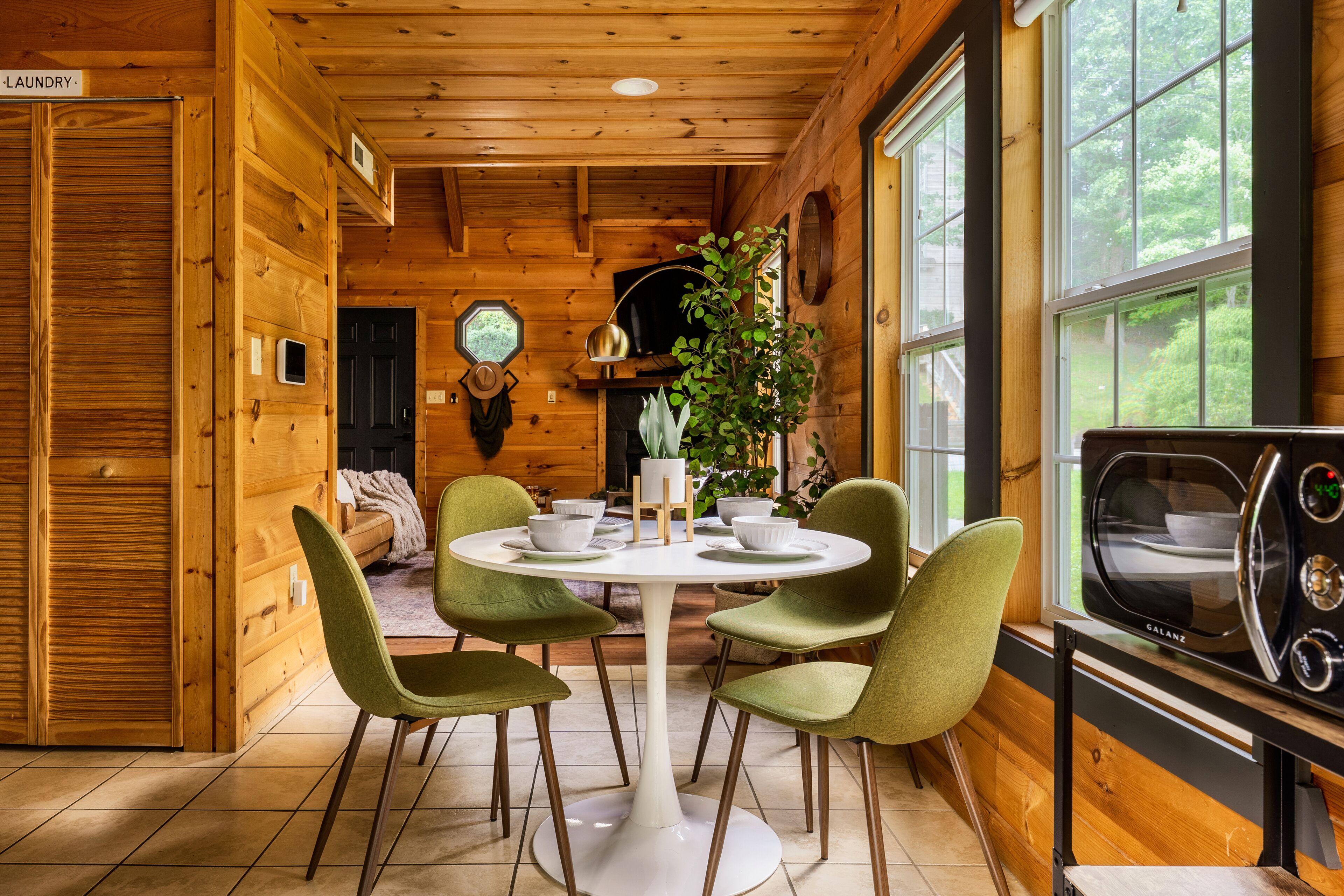 A wooden cabin interior with a dining table, four chairs, a potted plant, a clock on the wall, and a vintage radio.