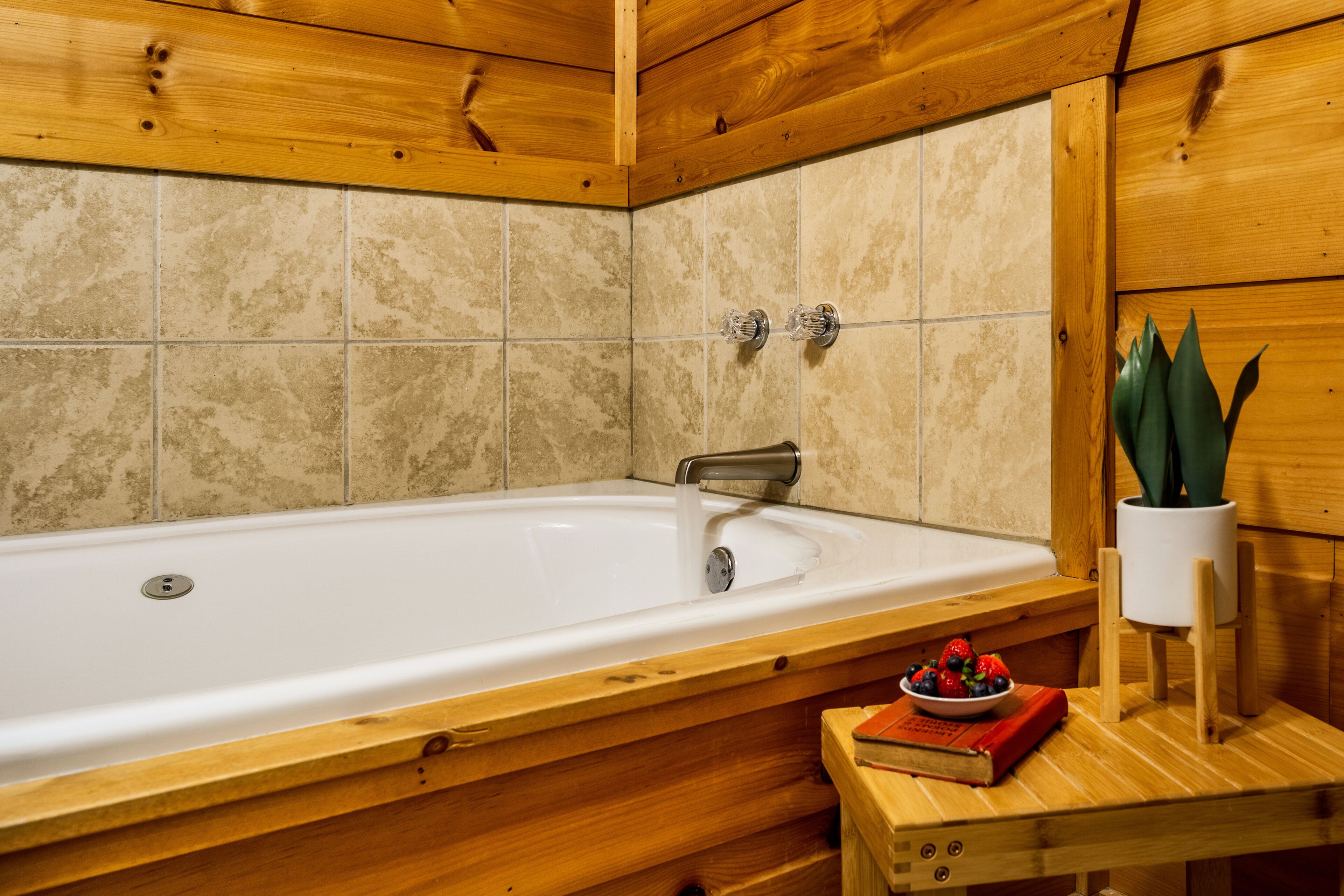 Une salle de bain rustique, avec des murs en bois, comprenant une baignoire, une petite table en bois sur laquelle est posé un saladier rempli de fruits, et une plante en pot.
