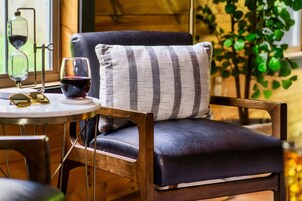 Uma cadeira com almofada escura e travesseiro listrado, um copo de vinho tinto e um par de óculos de sol sobre uma mesa pequena.