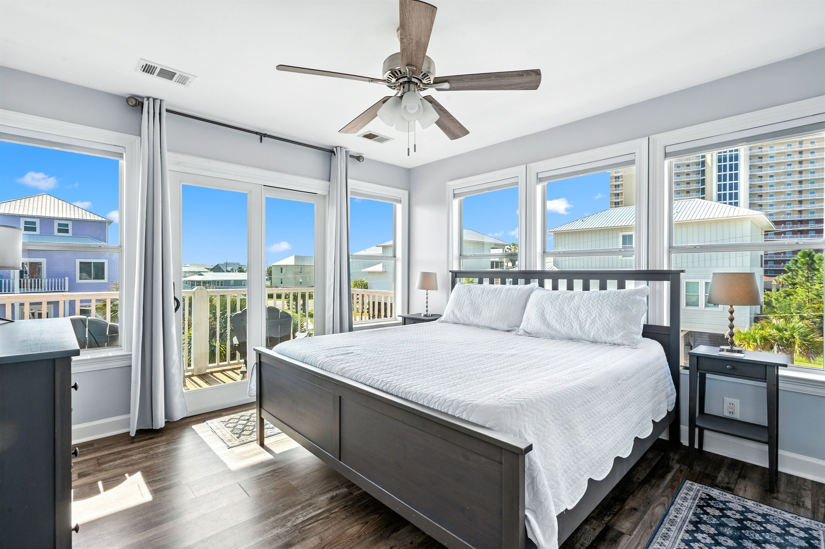 A bedroom with a large bed, a ceiling fan, and a view of neighboring houses through large windows.