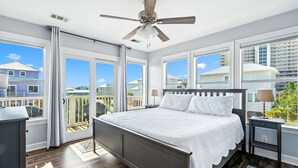 Un dormitorio con una cama grande, un ventilador de techo y vistas a casas vecinas a través de amplios ventanales.