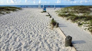 Un sendero arenoso con postes de madera que lleva a una zona de playa con sombrillas y gente a lo lejos.