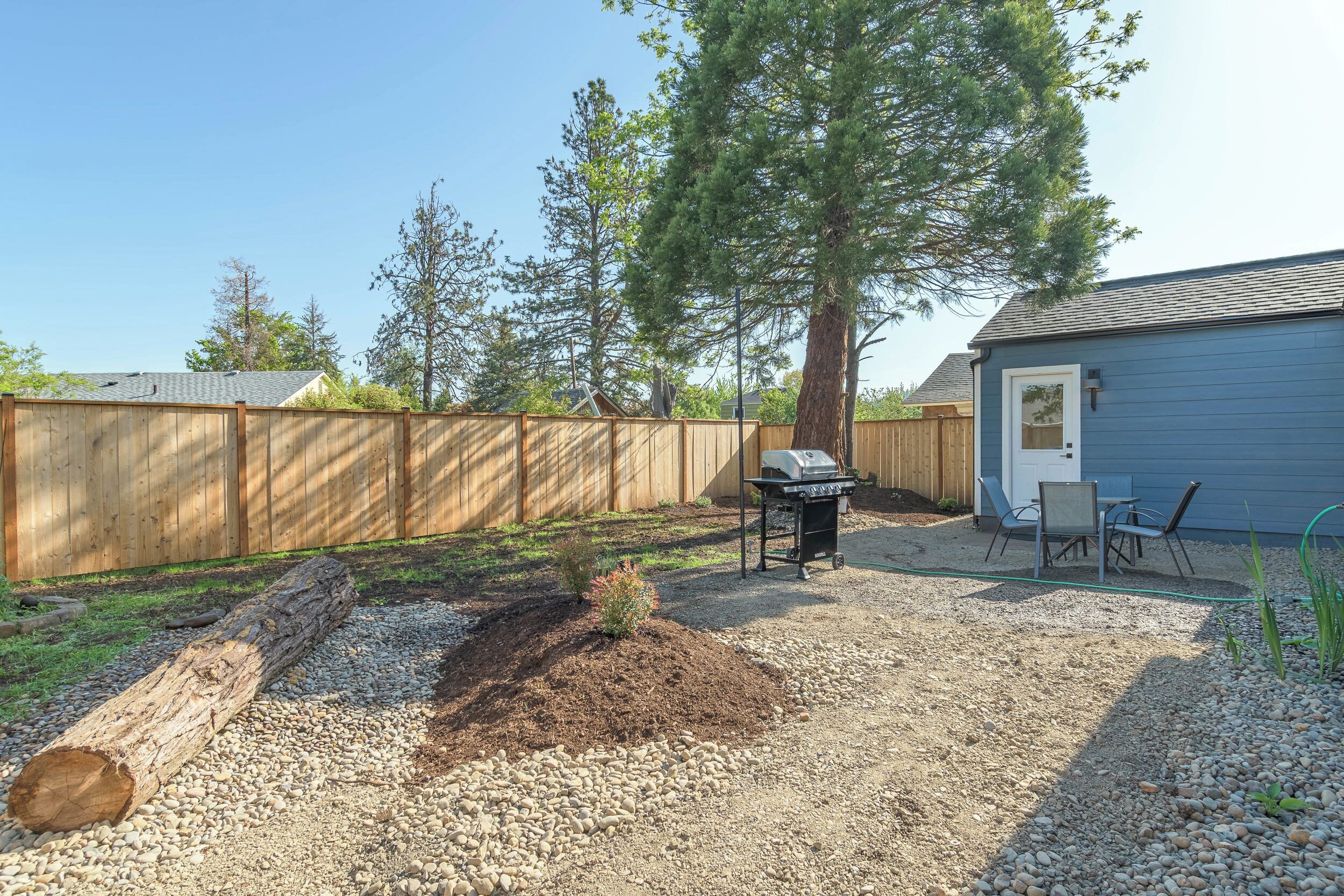 Un jardin avec une clôture en bois, un barbecue et un petit espace patio aménagé avec des chaises.