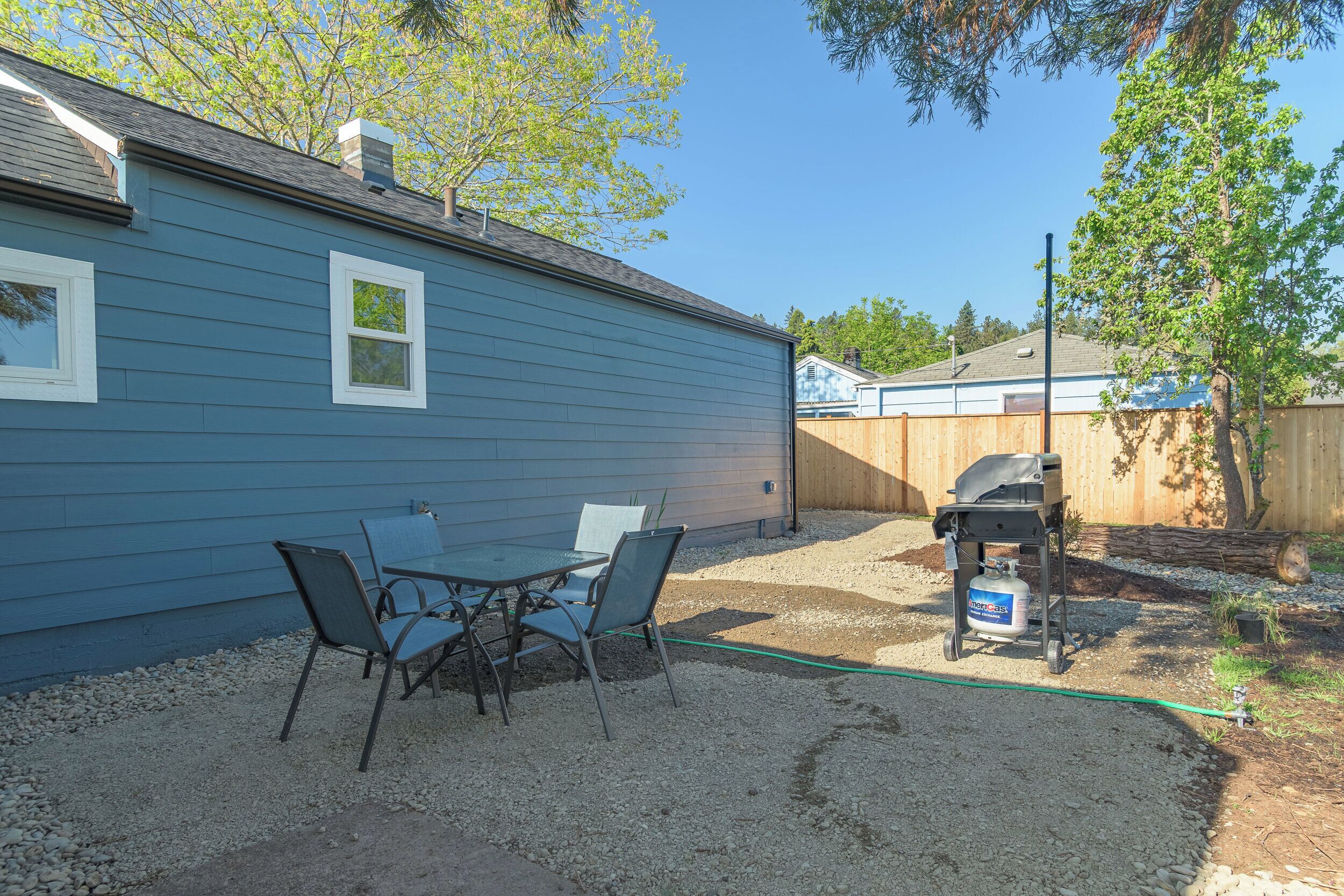 A backyard with a blue house, a patio table with chairs, a grill, and a propane tank.