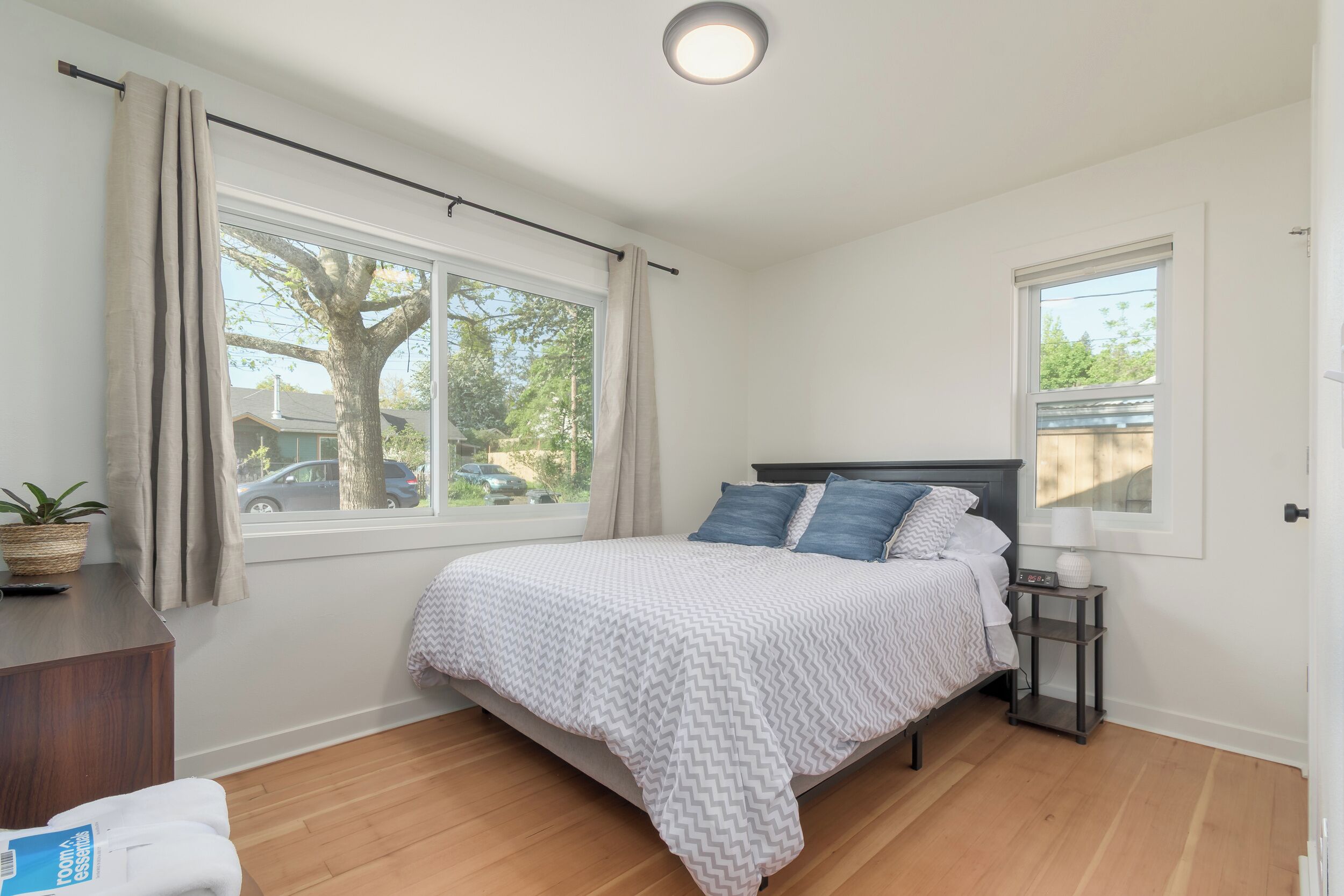 Une chambre à coucher avec un lit, une table de chevet, une fenêtre avec des rideaux, une plante et une vue sur l’extérieur.