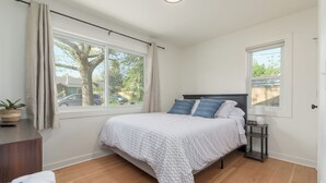 Une chambre à coucher avec un lit, une table de chevet, une fenêtre avec des rideaux, une plante et une vue sur l’extérieur.