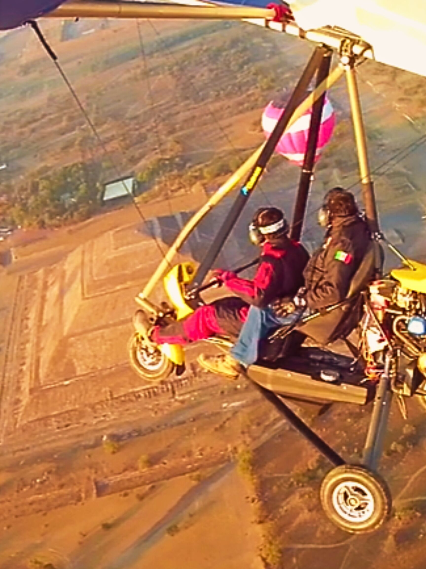 Eine Person in roter Jacke und Helm sitzt in einem Gleitschirm mit gelber Plane und fliegt über eine Landschaft mit Feldern und einem kleinen Gebäude.