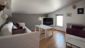 A modern living room with a white sofa, a white coffee table, a flat-screen TV, and a window providing natural light. - Furnished apartment with swimming pool (Saint-Georges-d'Oléron)