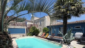 A rectangular swimming pool with a stone border, surrounded by palm trees and a blue building. - Furnished apartment with swimming pool (Saint-Georges-d'Oléron)