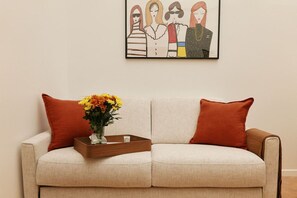 A beige sofa with two orange cushions, a wooden tray with a vase of yellow flowers, and a framed artwork above.