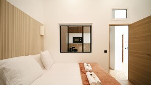 Ett modernt hotellrum med en stor säng, ett litet bord med handdukar, en inbyggd mikrovågsugn och utsikt över köket genom ett fönster.