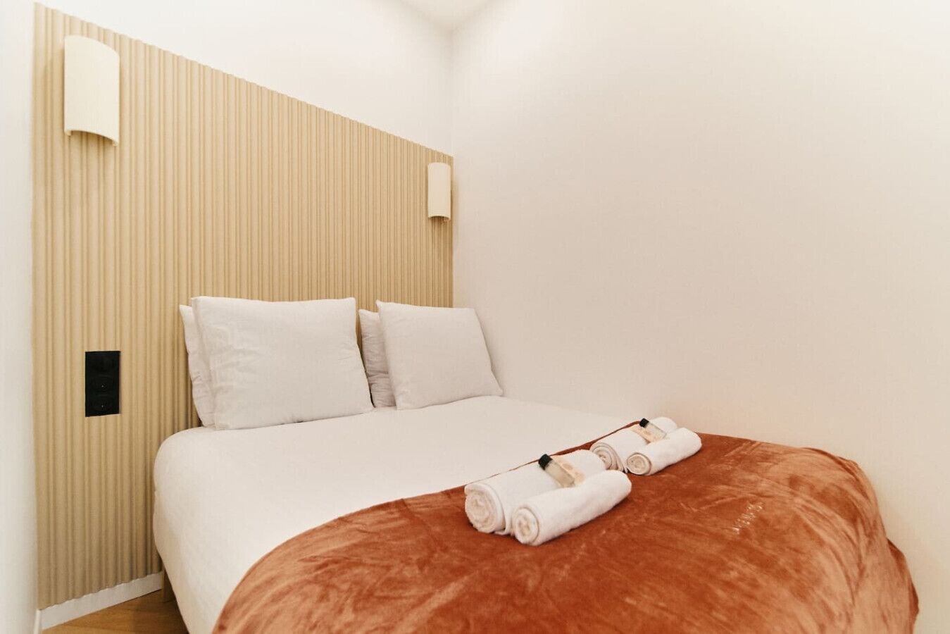 A hotel room with a bed, pillows, a headboard, a wall-mounted light, and a neatly folded towel on the bed.