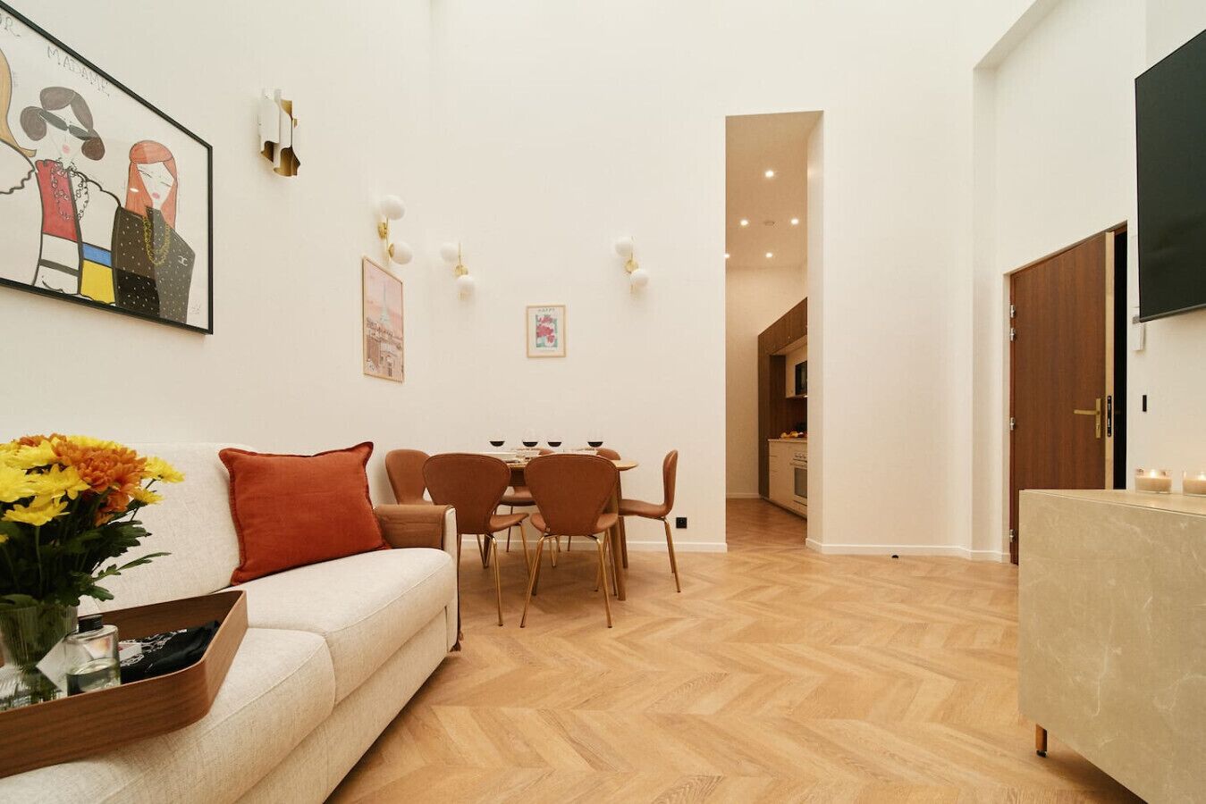 Ein modernes Wohnzimmer mit einer Couch, einem Esstisch mit Stühlen, einem an der Wand befestigten Fernseher und einer durch eine offene Tür sichtbaren Küche.
