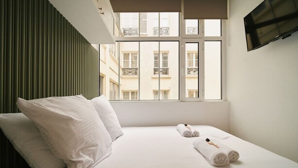 A hotel room with a bed, pillows, a window with a view of buildings, and a flat-screen TV mounted on the wall.