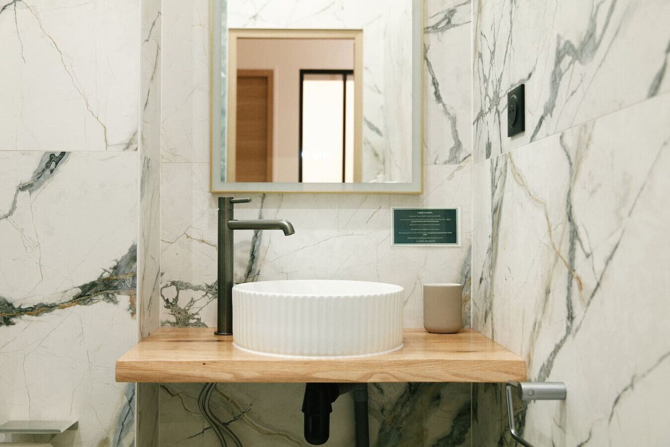 A bathroom with a marble wall, a round white sink, a wooden countertop, and a mirror above the sink.