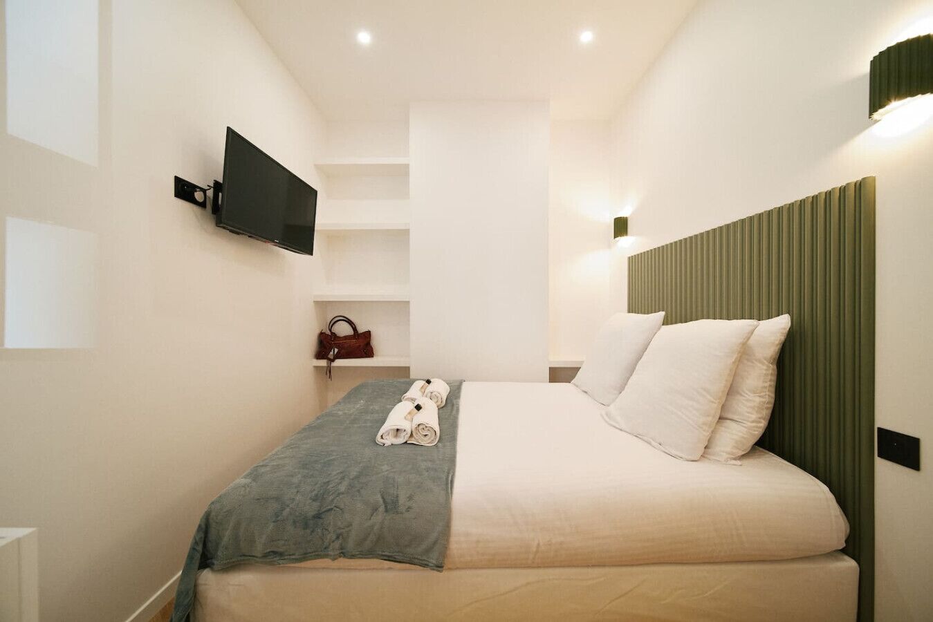 A modern hotel room with a bed, a flat-screen TV mounted on the wall, a small shelf with a handbag, and a green headboard.