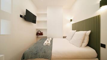 A modern hotel room with a bed, a flat-screen TV mounted on the wall, a small shelf with a handbag, and a green headboard.