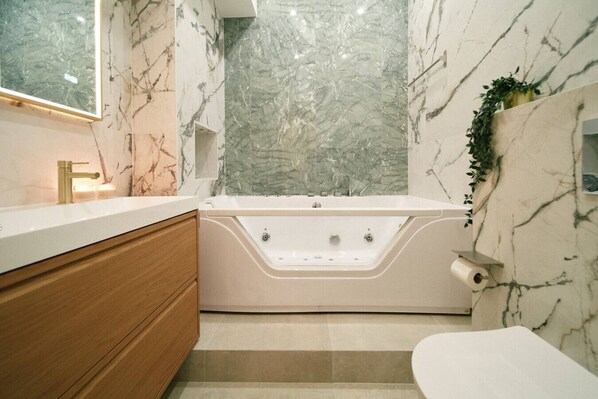 A modern bathroom with a marble wall, a bathtub, a wooden vanity, and a toilet.