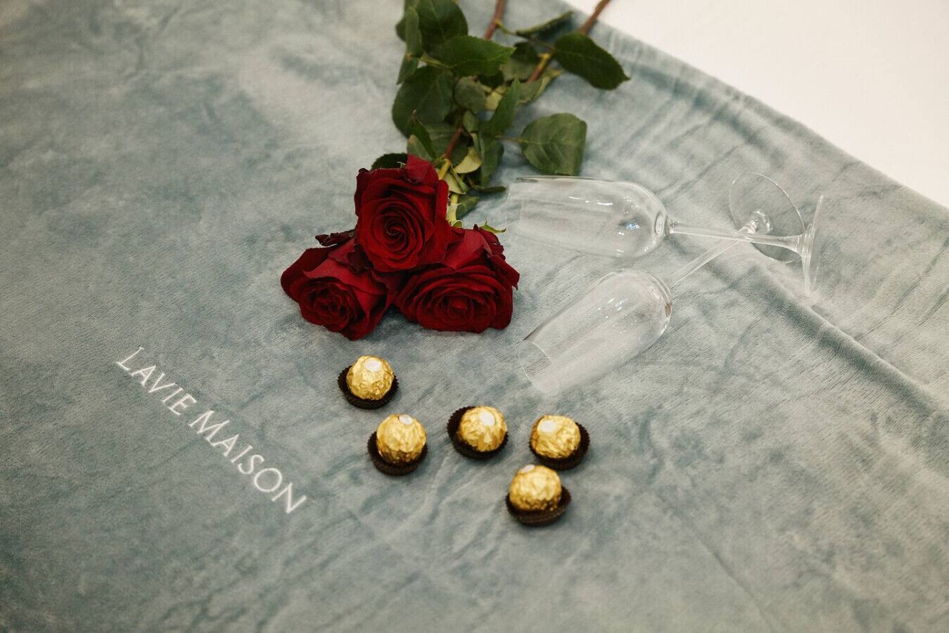 A grey fabric surface with a glass wine glass, a bouquet of red roses, and several wrapped chocolates.