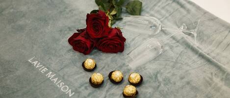 Eine graue Stoffoberfläche mit einem Weinglas, einem Rosenstrauß in Rot und mehreren verpackten Pralinen.