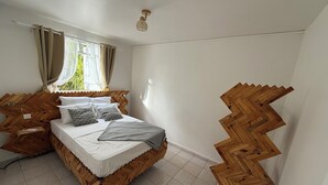 A bedroom with a wooden headboard, a bed with white linens and patterned pillows, a window with curtains, and a light fixture. - Villa apartment with private garden – quiet – 4 guests – Sainte-Anne (Sainte-Anne)