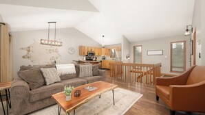 Una sala de estar moderna con un sofá, una mesa de centro y un sillón. Se ve una cocina al fondo, con armarios de madera y un comedor.