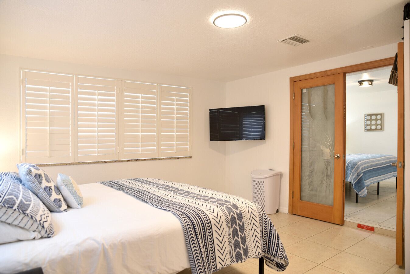 A bedroom with a bed, a television, a window with blinds, a door, and a tiled floor.