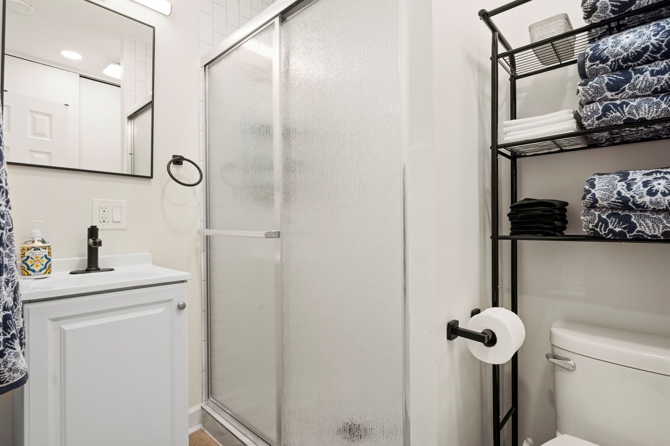Une salle de bain avec un lavabo blanc, une douche en verre, des toilettes et des étagères où sont pliées des serviettes.