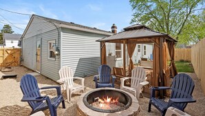 Ein Garten mit Feuerstelle, vier Adirondack-Stühlen und einem hölzernen Pavillon.