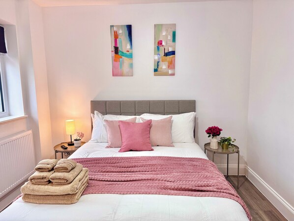 Ein ordentlich bezogenes Bett mit einer rosa Decke und einem Stapel beiger Handtücher. An der Wand über dem Bett hängen zwei abstrakte Gemälde.