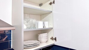 A kitchen cabinet with white shelves, holding clear glassware, white mugs, and stacked white plates. - Comfy Studio 30min to Central London (Wembley)