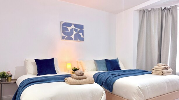 A hotel room with two beds, a wall-mounted artwork, a bedside lamp, and a window with curtains. - Comfy Studio 30min to Central London (Wembley)