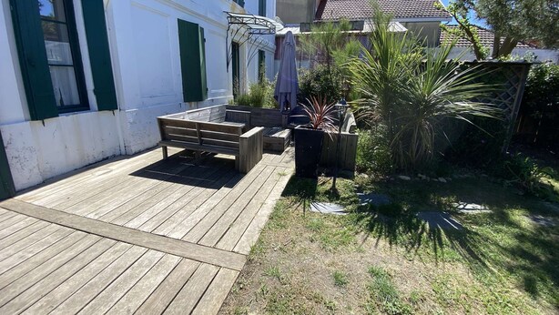 A wooden deck with a bench, a small garden with plants, and a white building with green shutters.