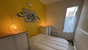 A bedroom with a bed, a nightstand, a wall-mounted light, a radiator, and a window with a view of a building.