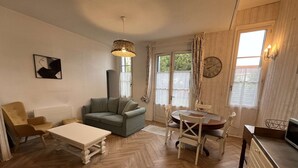 A cozy living room with a sofa, a dining table, and chairs. There is a large window with curtains, a clock on the wall, and a hanging light fixture.