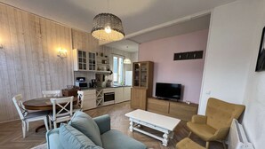 A modern living room with a sofa, a coffee table, and a TV. There is a kitchen area with cabinets and a dining table.