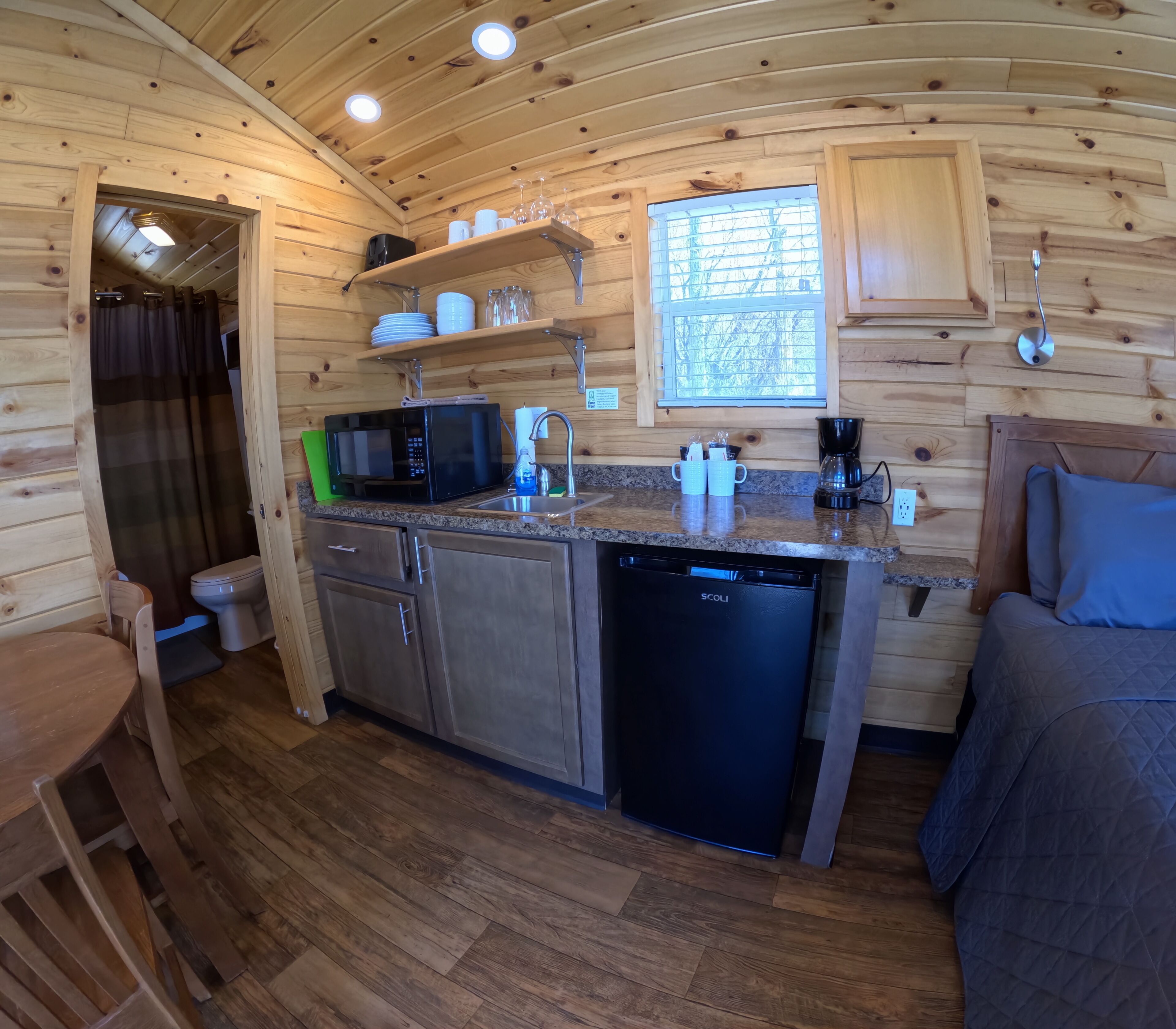Cocina compacta con microondas, cafetera y lavabo. Hay una ventana con persianas, un armario de madera y un pequeño comedor con mesa y sillas.