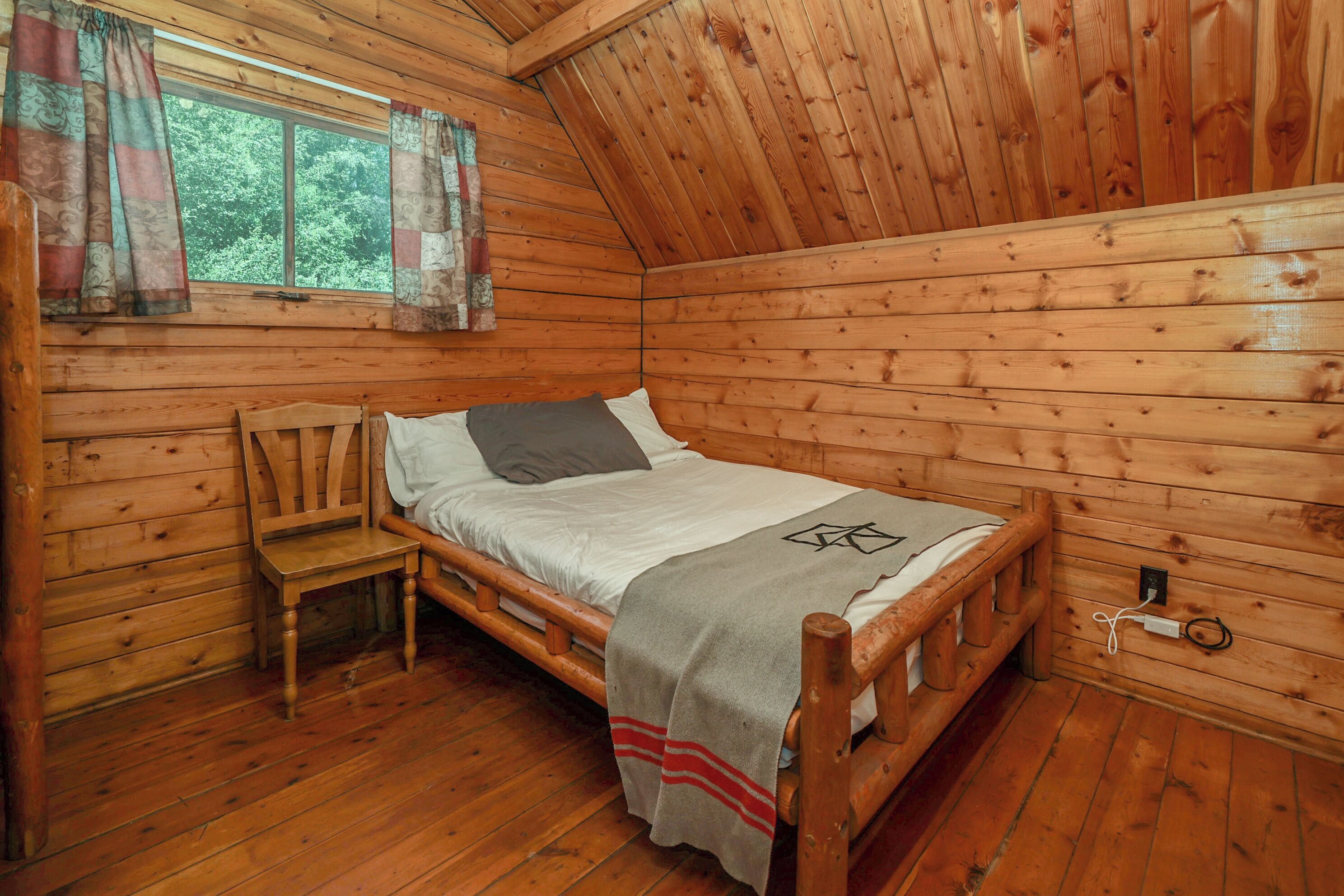 A wooden cabin room with a bed, a chair, a window with curtains, and a wooden floor.