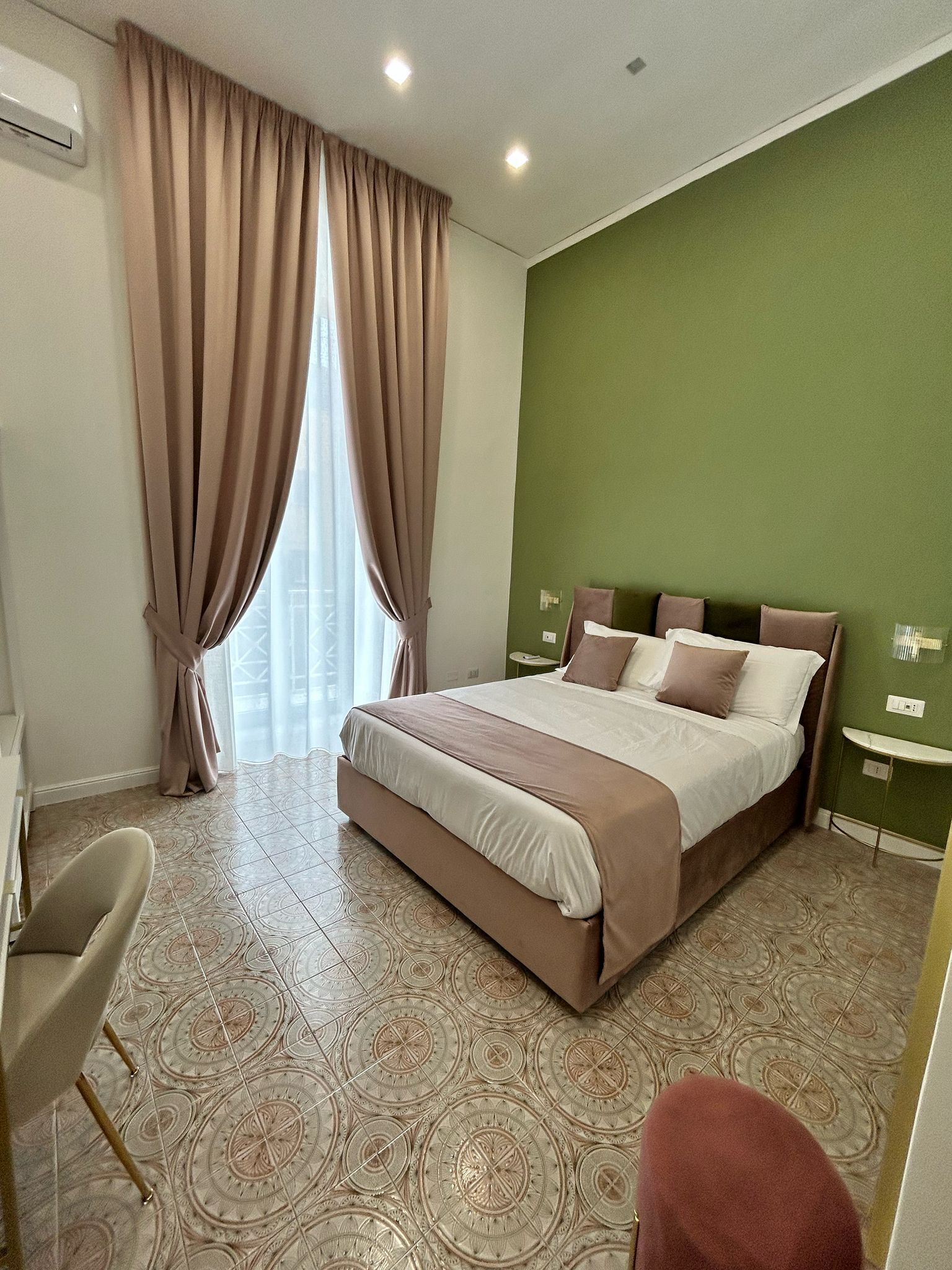 A hotel room with a bed, curtains, a chair, a small table, and a green wall.
