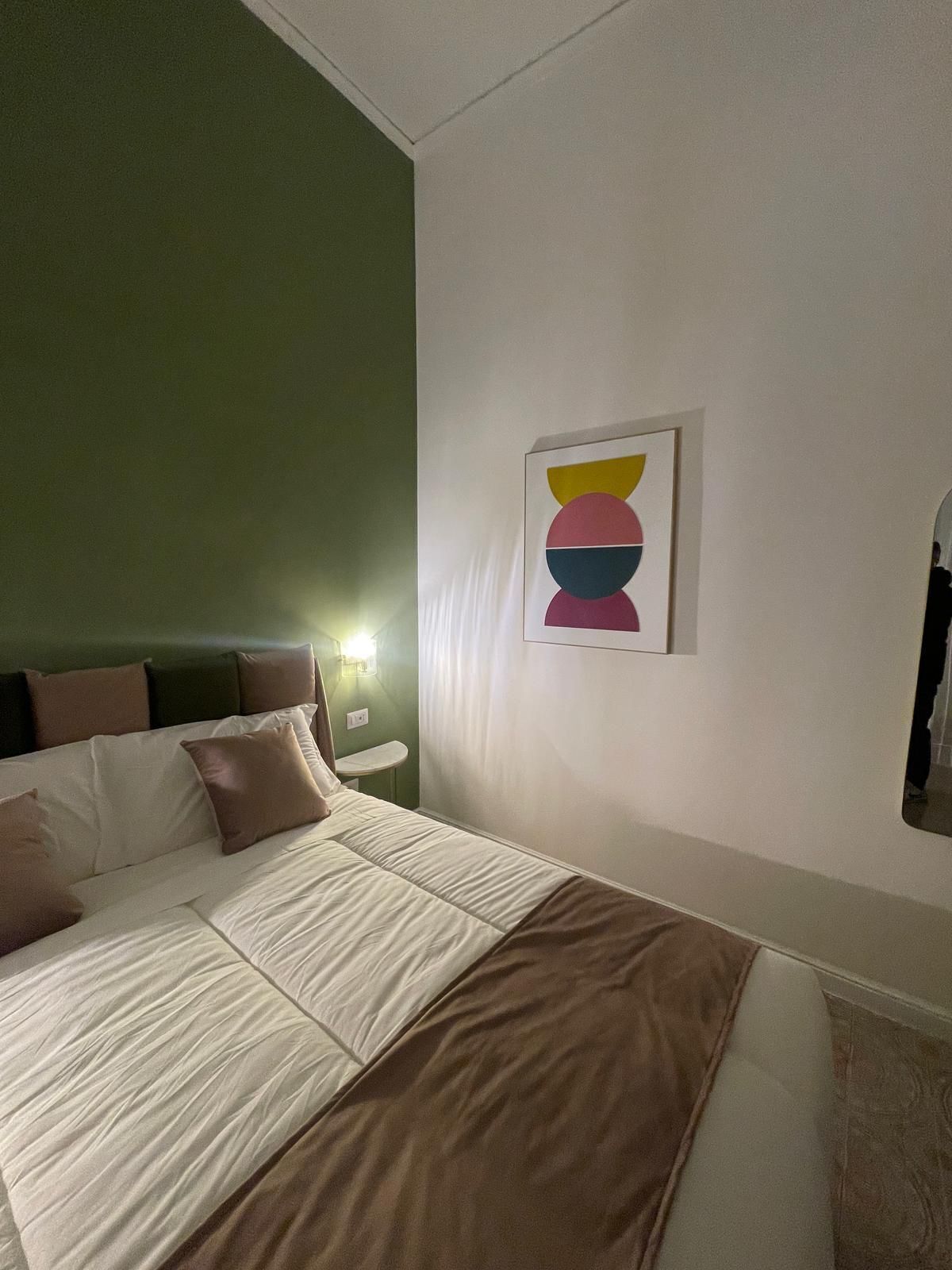 A neatly made bed with a brown blanket, a green wall, and a framed abstract art piece above it.