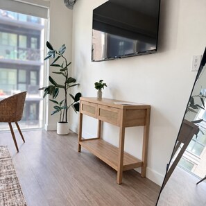 Ein Zimmer mit einem Holzkonsolentisch, einer Topfpflanze, einem Korbstuhl und einem an der Wand montierten Flachbildfernseher.