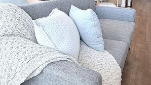 Un salon moderne avec un canapé gris, un plaid blanc en tricot et une cuisine en arrière-plan.