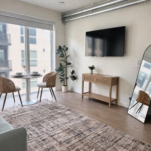 A modern living room with a large window, a flat-screen TV mounted on the wall, a wooden console table, and a large mirror reflecting the room. - Modern Downtown SLC | King Bed+Pool+Free Parking (Salt Lake City)