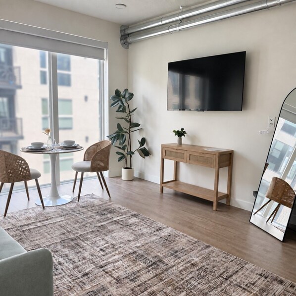A modern living room with a large window, a flat-screen TV mounted on the wall, a wooden console table, and a large mirror reflecting the room.
