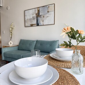 A dining area with a white table set for two, a blue sofa in the background, and a wall-mounted abstract painting.