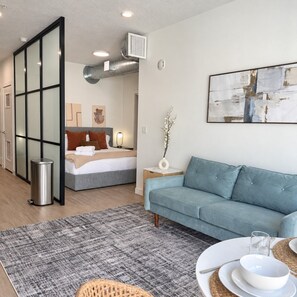A modern hotel room with a bed, a sofa, a dining table with plates and glasses, a trash bin, and a decorative vase.